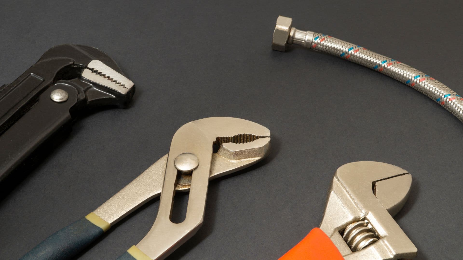 Plumbing tools including pipe wrenches, multigrips and a flexible supply hose laid out on a dark surface